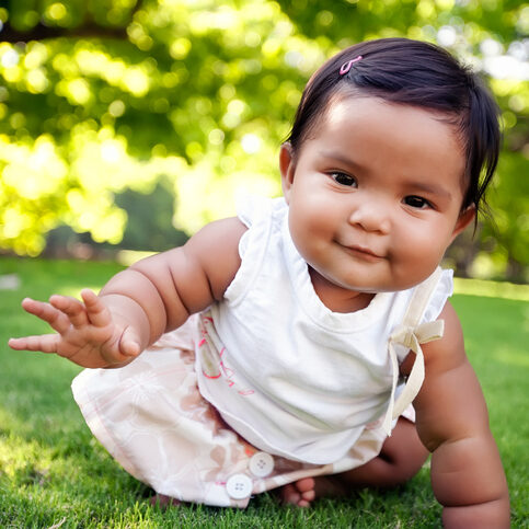 Cute baby girl with a smile on her face, reaching out to take her first crawling step on a lush green lawn at an outdoor park, of mixed ethnic race.